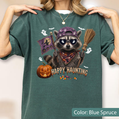 May include: A blue spruce t-shirt featuring a graphic of a raccoon wearing a purple cap, heart-shaped sunglasses, and a patterned bandana. The raccoon holds a broom and a flag with a witch silhouette. Text reads HAPPY HAUNTING with pumpkins.