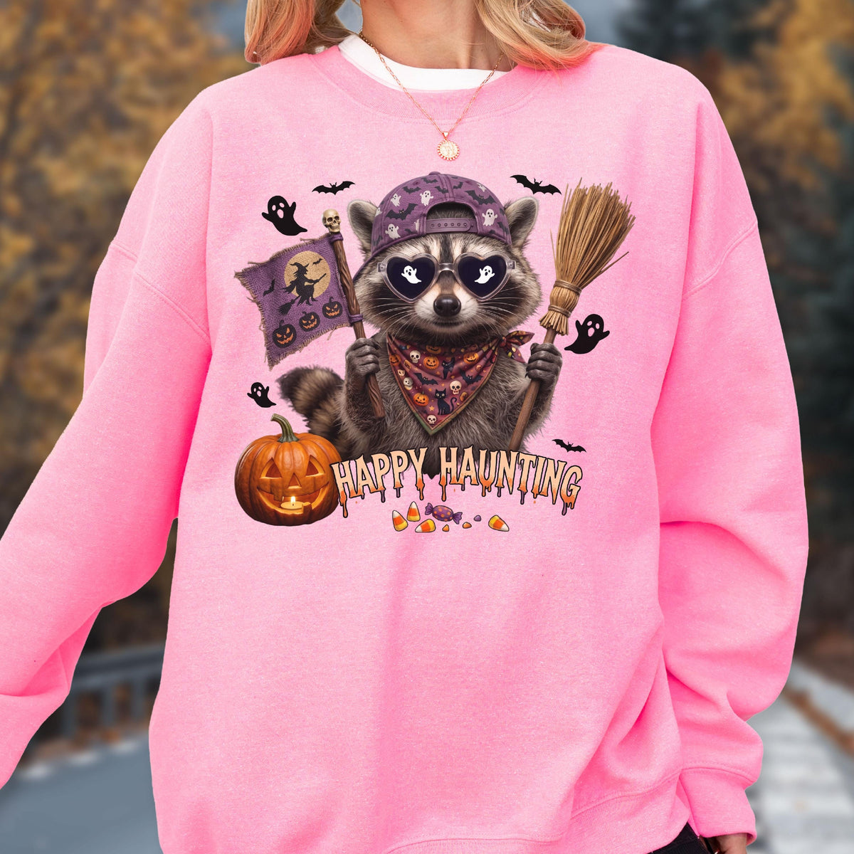 May include: A bright pink crewneck sweatshirt featuring a graphic of a raccoon wearing a purple baseball cap, heart-shaped sunglasses, and a patterned bandana. The raccoon holds a broom and a flag with a witch silhouette. Text reads HAPPY HAUNTING.