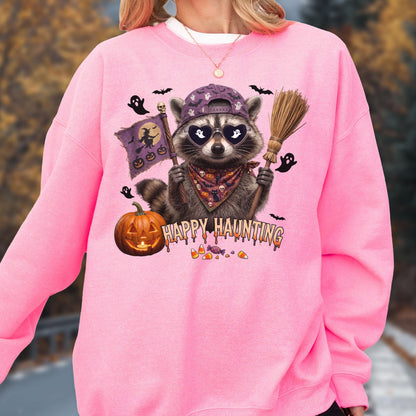 May include: A bright pink crewneck sweatshirt featuring a graphic of a raccoon wearing a purple baseball cap, heart-shaped sunglasses, and a patterned bandana. The raccoon holds a broom and a flag with a witch silhouette. Text reads HAPPY HAUNTING.