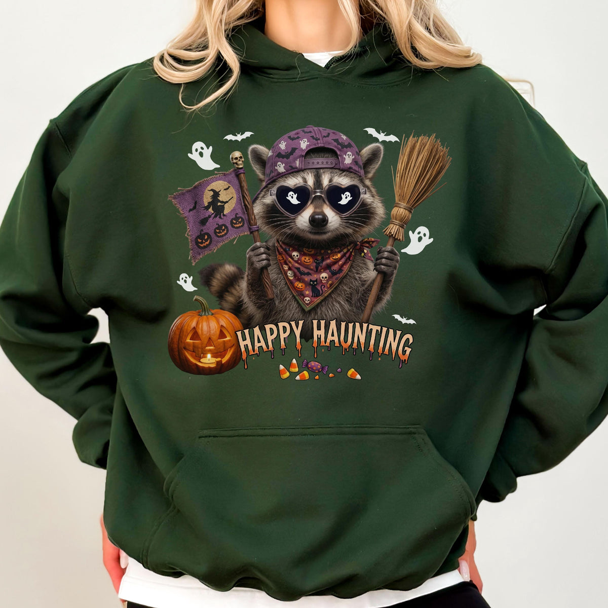 May include: A dark green hooded sweatshirt featuring a graphic of a raccoon wearing a purple baseball cap and heart-shaped sunglasses. The raccoon holds a broom and a flag with a witch silhouette. Text reads Happy Haunting with candy corn.