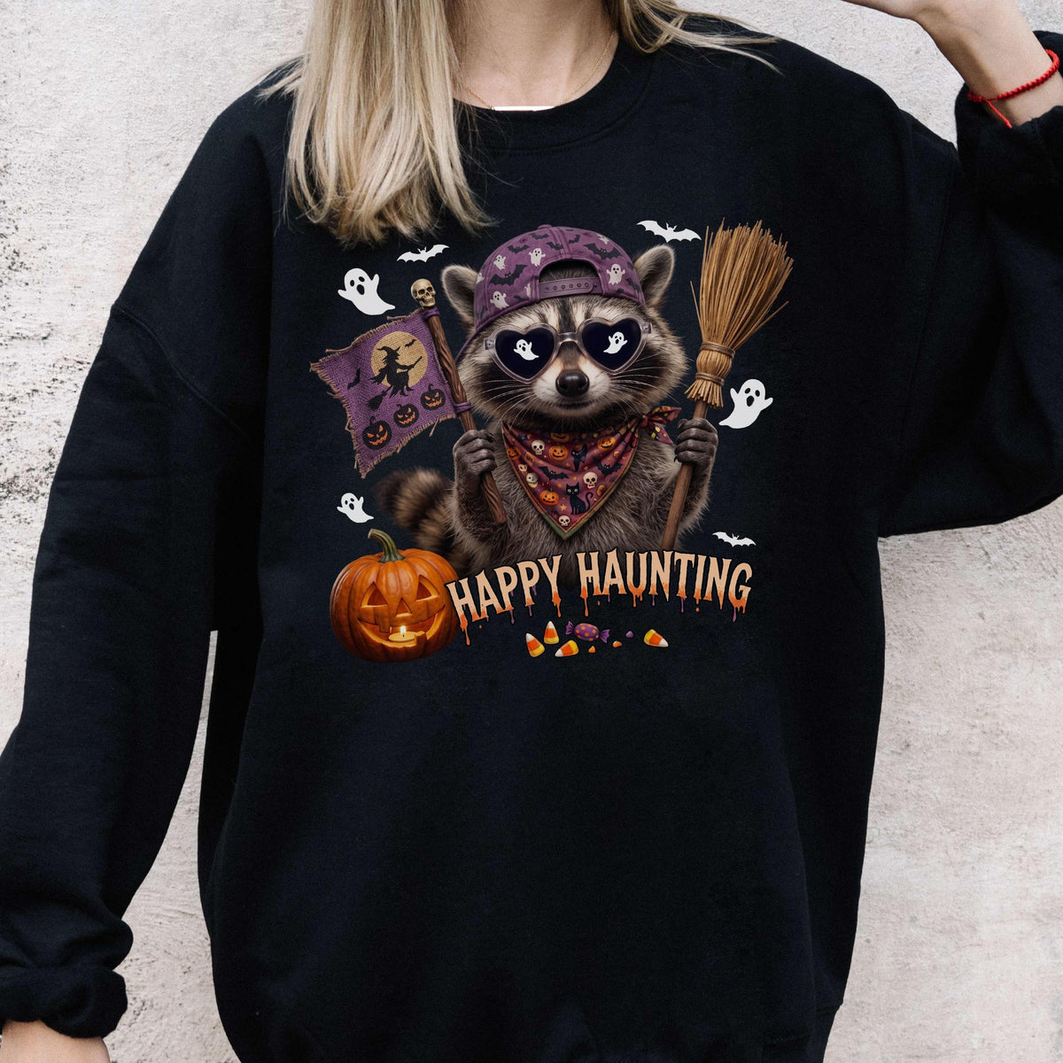 May include: A black sweatshirt featuring a graphic of a raccoon wearing a purple baseball cap, heart-shaped sunglasses, and a patterned bandana. The raccoon holds a broom and a flag with a witch and pumpkins. Text reads HAPPY HAUNTING.