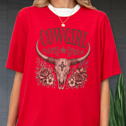 May include: A bright red short-sleeve t-shirt featuring a graphic print of a longhorn skull centered on the chest. The word COWGIRL is printed in large dark letters above the skull, which is flanked by detailed illustrations of roses.