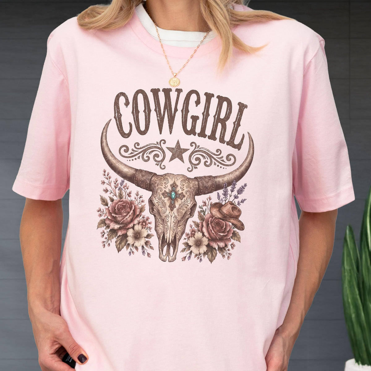 May include: A light pink t-shirt featuring a graphic print of a longhorn skull with a central turquoise stone, surrounded by roses, lavender, and a small cowboy hat. The word COWGIRL is printed in brown above the skull with a star and scrollwork.