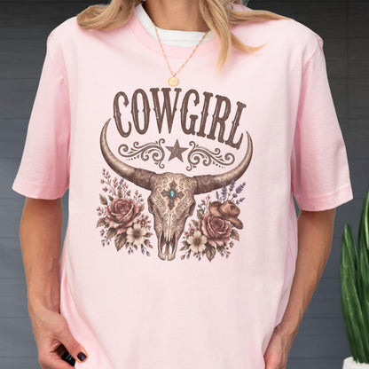 May include: A light pink t-shirt featuring a graphic print of a longhorn skull with a central turquoise stone, surrounded by roses, lavender, and a small cowboy hat. The word COWGIRL is printed in brown above the skull with a star and scrollwork.