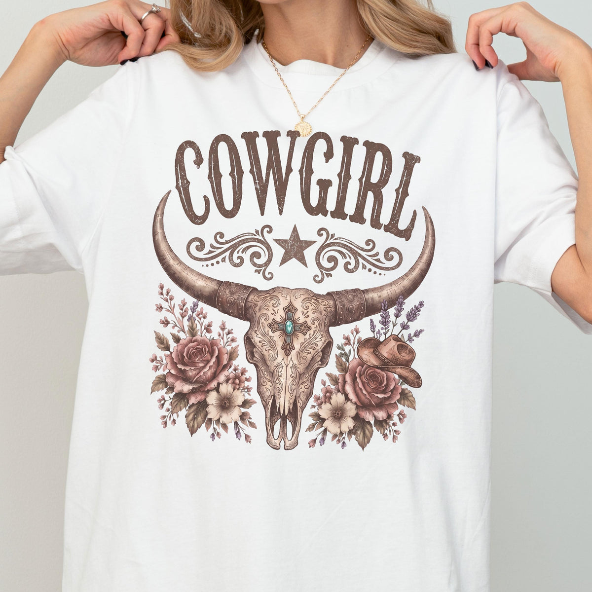 May include: A white t-shirt featuring a brown graphic of a longhorn skull with a turquoise center accent, surrounded by roses, lavender, and a small cowboy hat. The word COWGIRL is printed in brown above the skull with a star and scroll design.