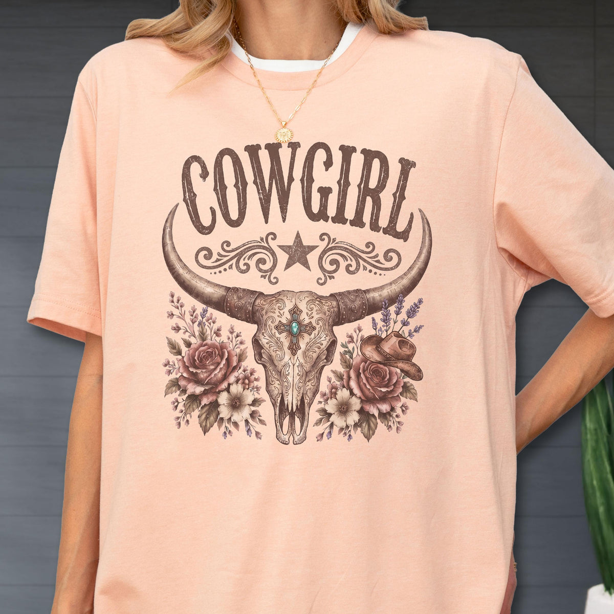 May include: A peach-colored t-shirt featuring a graphic print of a longhorn skull centered below the text COWGIRL. The design includes a star, floral accents, and a small cowboy hat, all rendered in shades of brown, cream, and turquoise blue.