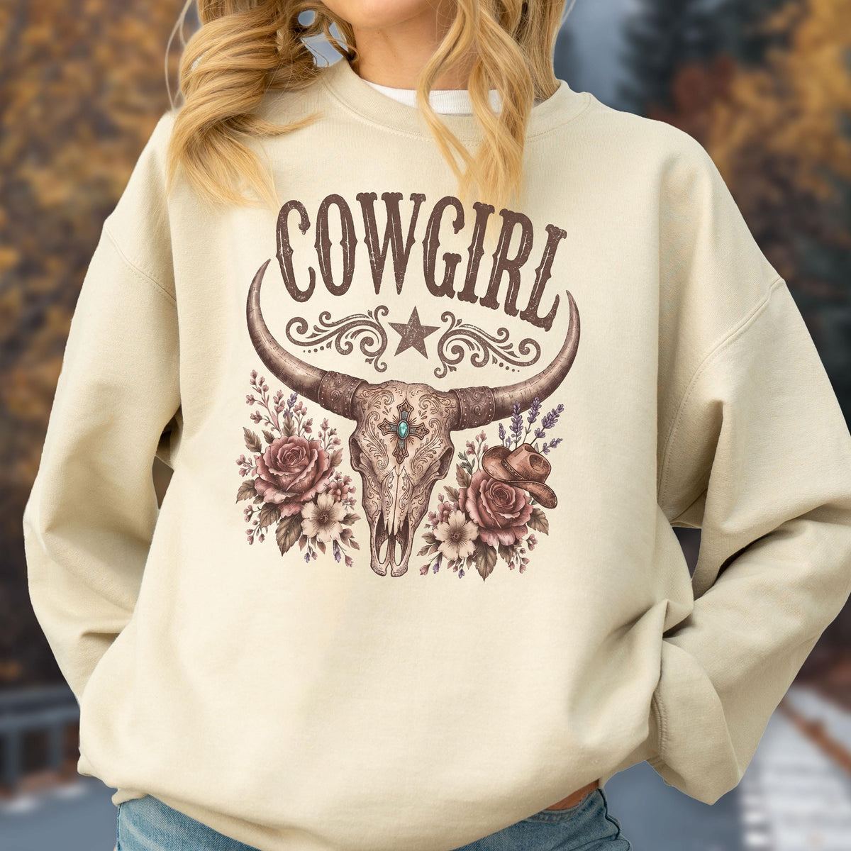 May include: A beige crewneck sweatshirt featuring a brown graphic print of a longhorn skull centered on the chest. The design includes the text COWGIRL above the skull, flanked by floral arrangements, a small star, and a cowboy hat illustration.