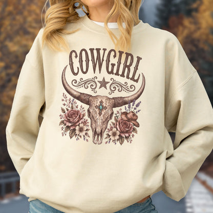 May include: A beige crewneck sweatshirt featuring a brown graphic print of a longhorn skull centered on the chest. The design includes the text COWGIRL above the skull, flanked by floral arrangements, a small star, and a cowboy hat illustration.