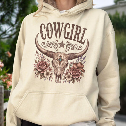 May include: A cream-colored hooded sweatshirt featuring a brown graphic print. The design displays the word COWGIRL above a steer skull with a central blue stone, surrounded by floral accents, a small cowboy hat, and a star motif.