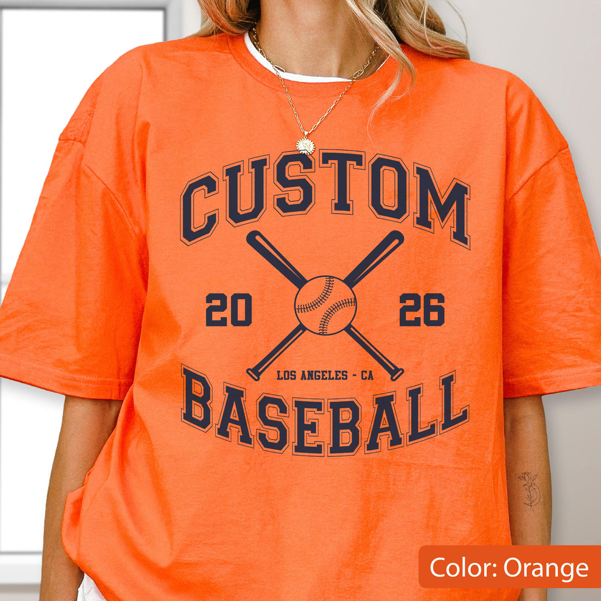 May include: An orange short-sleeve t-shirt featuring a dark blue graphic with the text CUSTOM BASEBALL, the numbers 20 and 26, and an illustration of a baseball with two crossed bats. The text LOS ANGELES - CA is printed below the baseball.