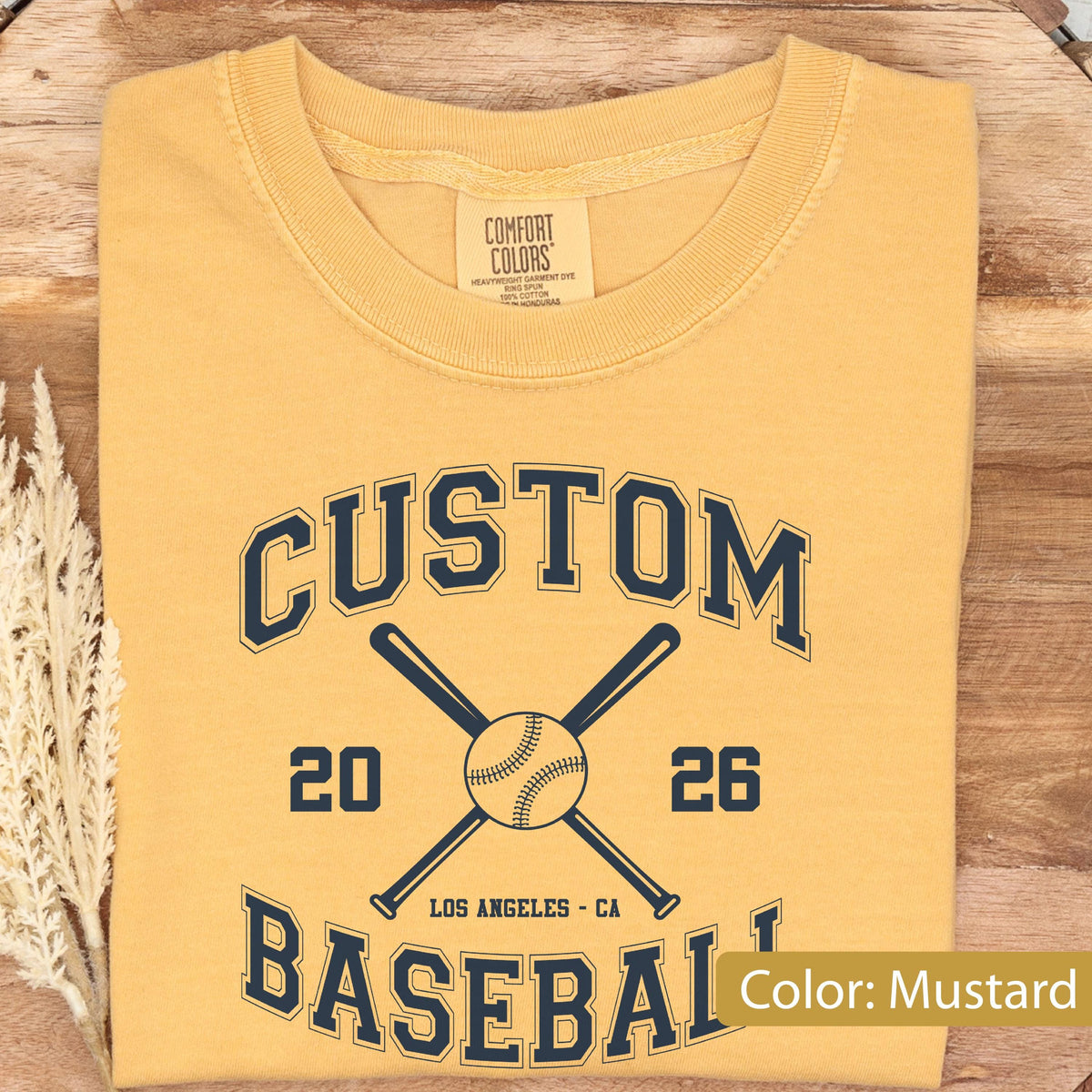 May include: A mustard yellow t-shirt featuring a dark navy blue graphic print. The design includes the text CUSTOM BASEBALL, the numbers 20 and 26, and the words LOS ANGELES CA, centered around an illustration of two crossed baseball bats and a ball.