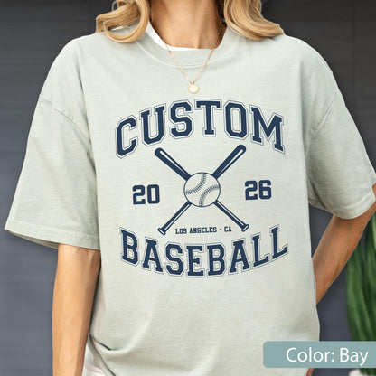 May include: A light sage green short-sleeve t-shirt featuring a dark navy blue graphic print. The design includes the text CUSTOM BASEBALL, the numbers 20 and 26, the words LOS ANGELES, and an illustration of a baseball with two crossed bats.