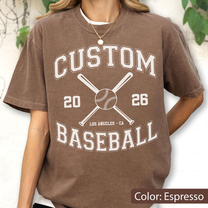 May include: An espresso brown short-sleeve t-shirt featuring a white graphic print with the text CUSTOM BASEBALL, the numbers 20 and 26, and the words LOS ANGELES - CA, centered around an illustration of a baseball and two crossed bats.