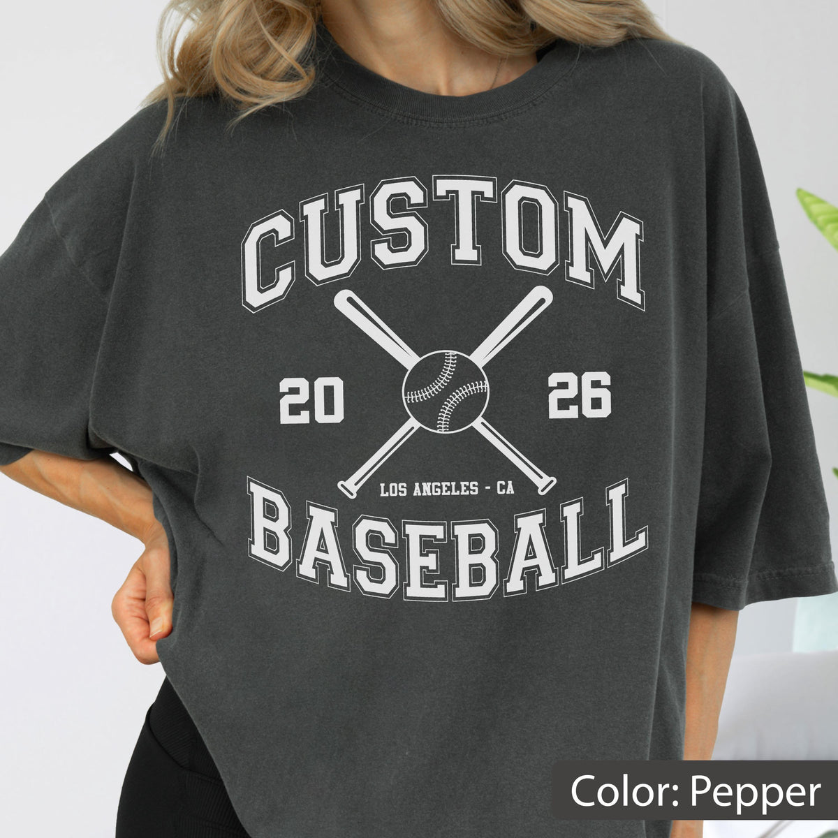 May include: A pepper-colored t-shirt featuring a white graphic print with the text CUSTOM BASEBALL, 20 26, and LOS ANGELES - CA. The design includes a central baseball illustration positioned over two crossed baseball bats in a collegiate font style.
