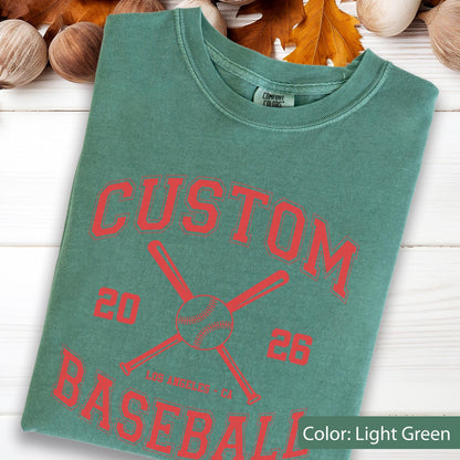 May include: A light green short-sleeve t-shirt featuring a red graphic print. The design includes the text CUSTOM BASEBALL, the numbers 20 and 26, and an illustration of two crossed baseball bats with a baseball. The shirt has a crew neckline.