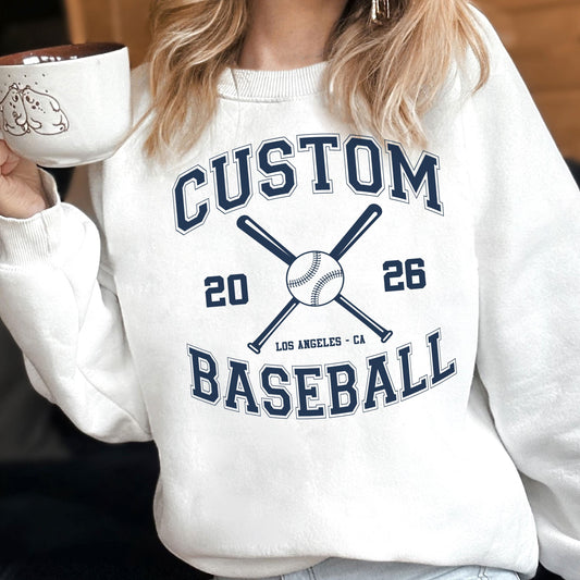 May include: A white crewneck sweatshirt featuring a dark blue graphic print. The design includes the text CUSTOM BASEBALL, the numbers 20 and 26, the location LOS ANGELES - CA, and an illustration of a baseball with two crossed baseball bats.