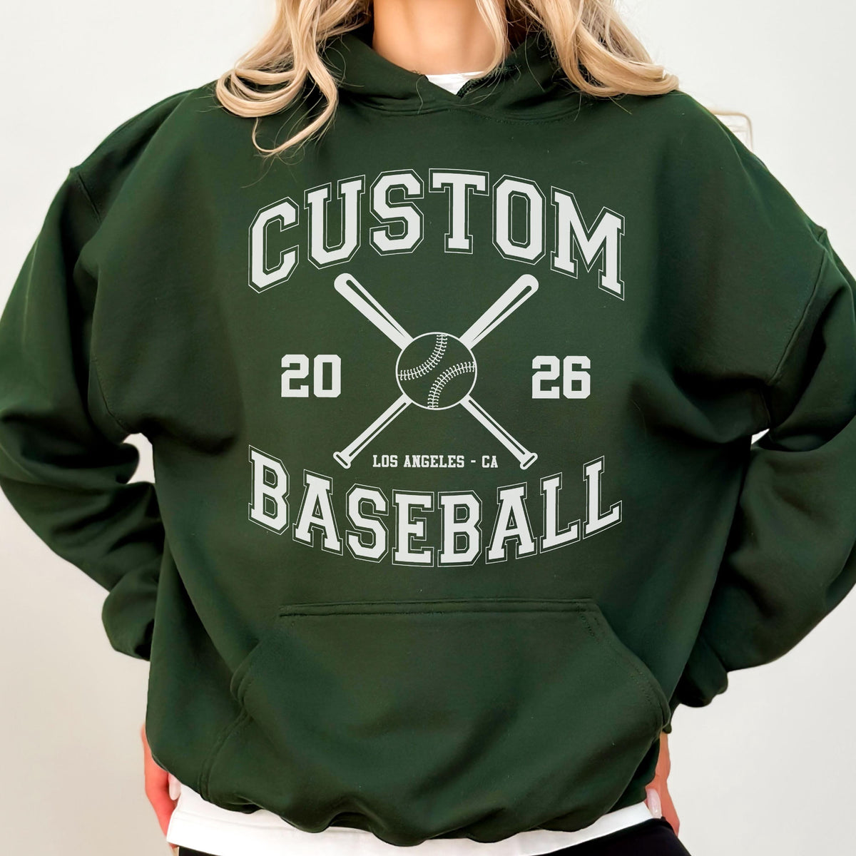 May include: A dark green hooded sweatshirt featuring a white graphic print with the text CUSTOM BASEBALL, the numbers 20 and 26, the location LOS ANGELES - CA, and an illustration of a baseball centered between two crossed baseball bats.