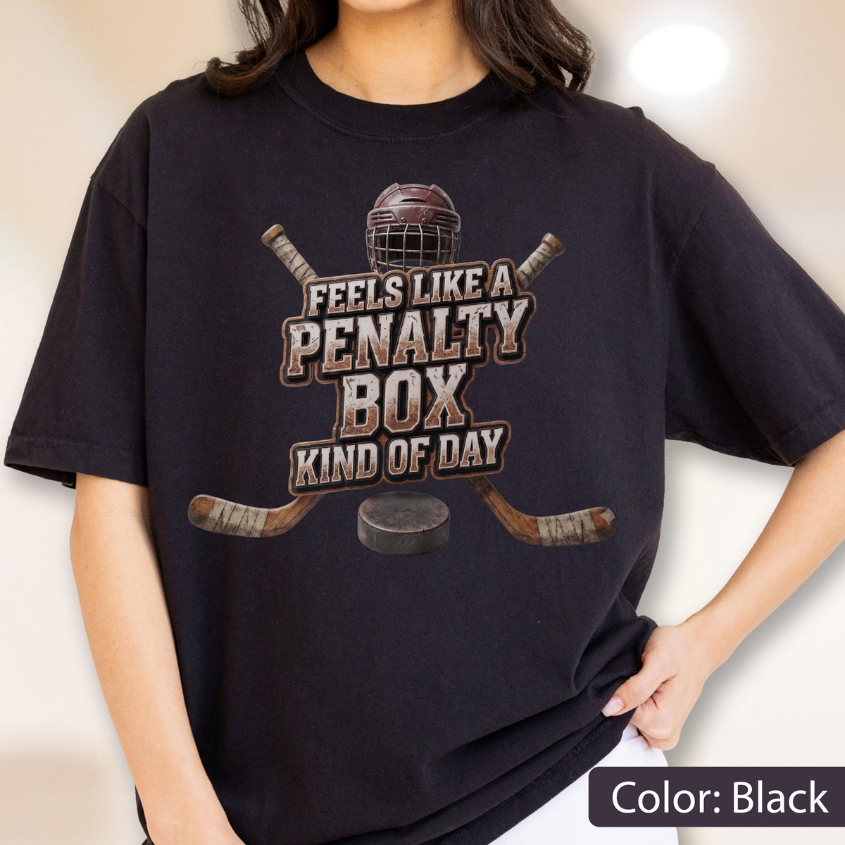 May include: A black short-sleeved t-shirt featuring a graphic of a hockey helmet, two crossed hockey sticks, and a puck. The text reads FEELS LIKE A PENALTY BOX KIND OF DAY in a distressed, weathered font style centered on the chest area.