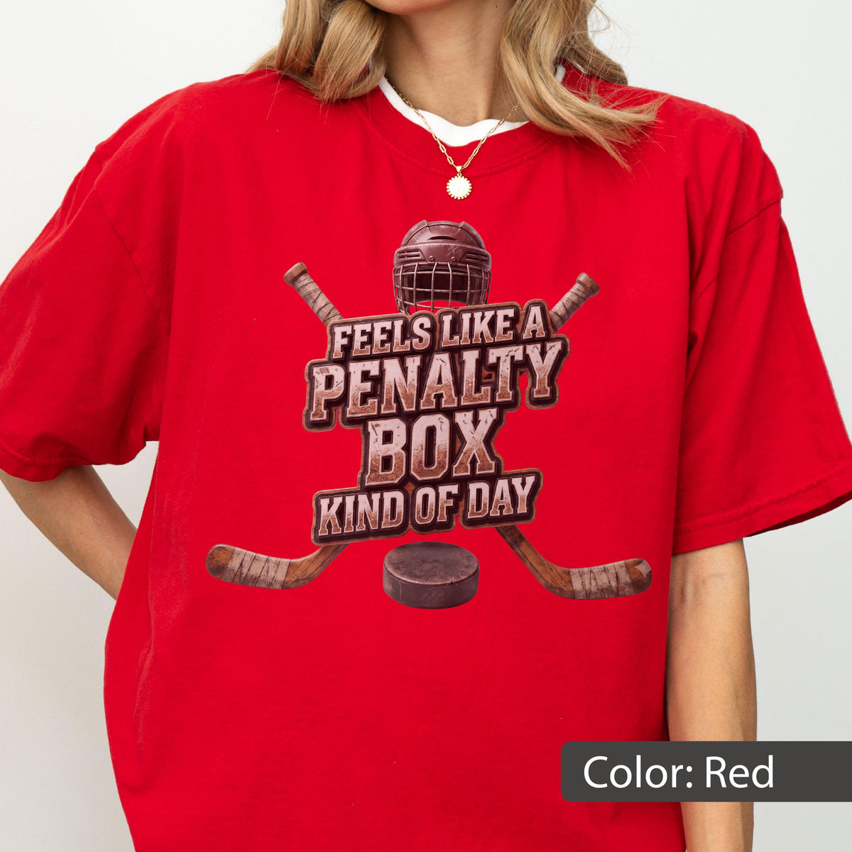 May include: A bright red short-sleeved t-shirt featuring a graphic of a hockey helmet, two crossed hockey sticks, and a puck. The text reads FEELS LIKE A PENALTY BOX KIND OF DAY in a distressed brown font centered on the chest of the garment.