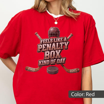 May include: A bright red short-sleeved t-shirt featuring a graphic of a hockey helmet, two crossed hockey sticks, and a puck. The text reads FEELS LIKE A PENALTY BOX KIND OF DAY in a distressed brown font centered on the chest of the garment.