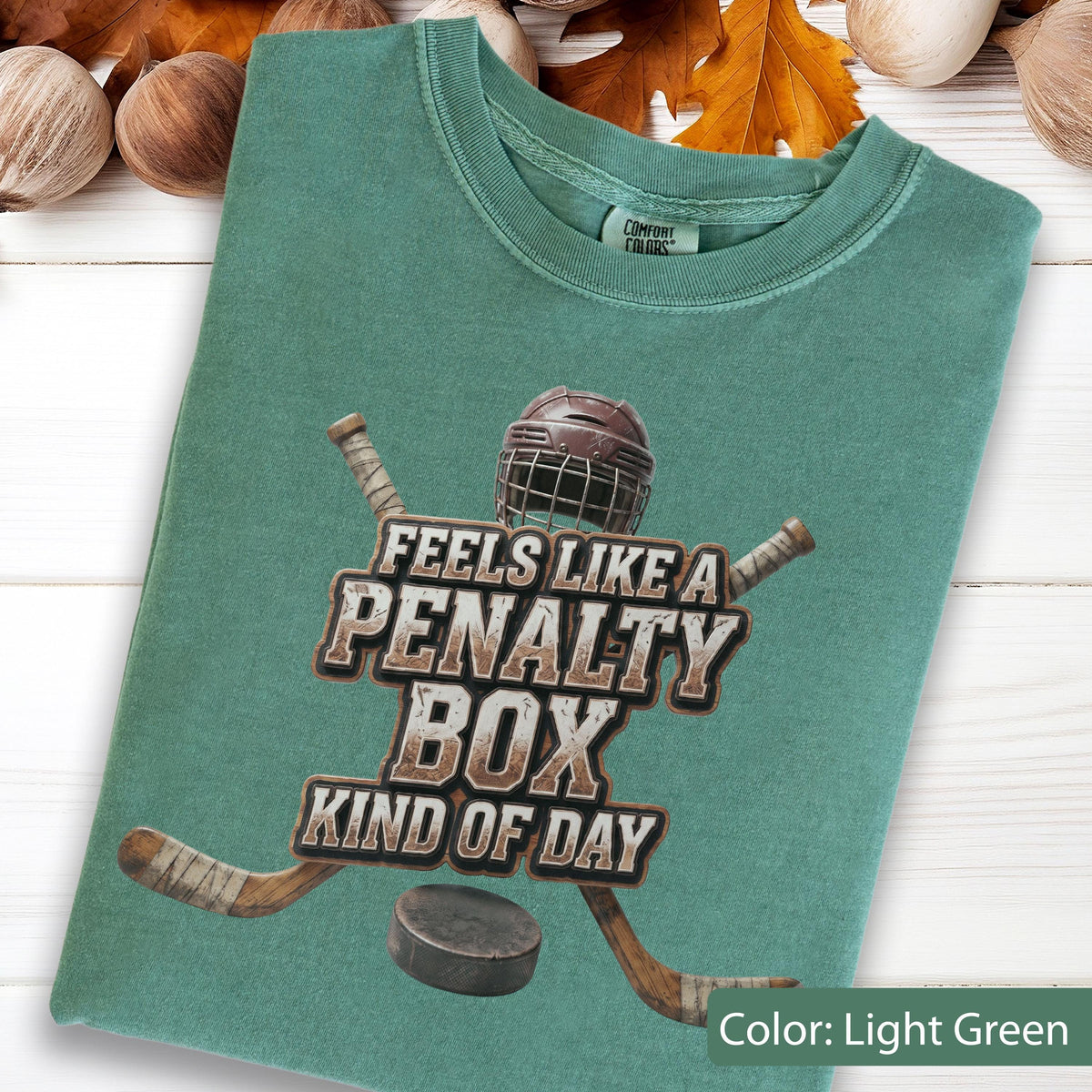 May include: A light green t-shirt featuring a graphic of a brown hockey helmet, two crossed hockey sticks, and a black puck. The text reads: FEELS LIKE A PENALTY BOX KIND OF DAY. The shirt has a crew neckline and a Comfort Colors brand label.