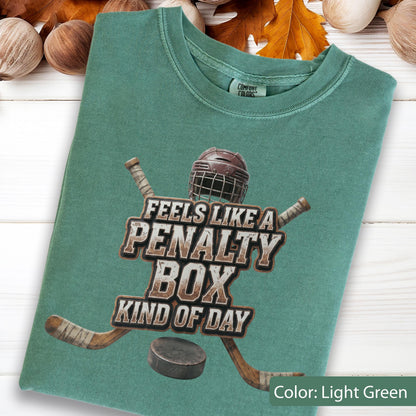 May include: A light green t-shirt featuring a graphic of a brown hockey helmet, two crossed hockey sticks, and a black puck. The text reads: FEELS LIKE A PENALTY BOX KIND OF DAY. The shirt has a crew neckline and a Comfort Colors brand label.