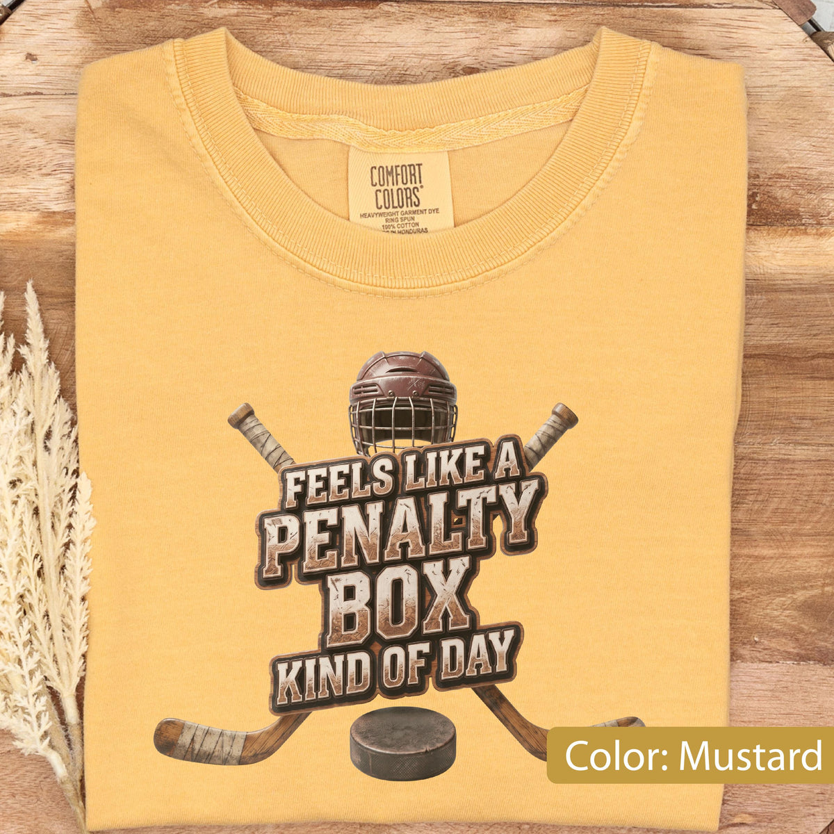 May include: A mustard yellow short-sleeve t-shirt with a graphic print featuring a hockey helmet, two crossed hockey sticks, and a hockey puck. The text reads "FEELS LIKE A PENALTY BOX KIND OF DAY". The shirt has a Comfort Colors brand neck label.