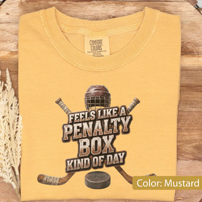 May include: A mustard yellow short-sleeve t-shirt with a graphic print featuring a hockey helmet, two crossed hockey sticks, and a hockey puck. The text reads "FEELS LIKE A PENALTY BOX KIND OF DAY". The shirt has a Comfort Colors brand neck label.