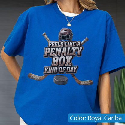 May include: A royal blue short-sleeve t-shirt featuring a graphic of a hockey helmet, two crossed hockey sticks, and a puck. The text reads, "FEELS LIKE A PENALTY BOX KIND OF DAY." The design is centered on the chest of the shirt.