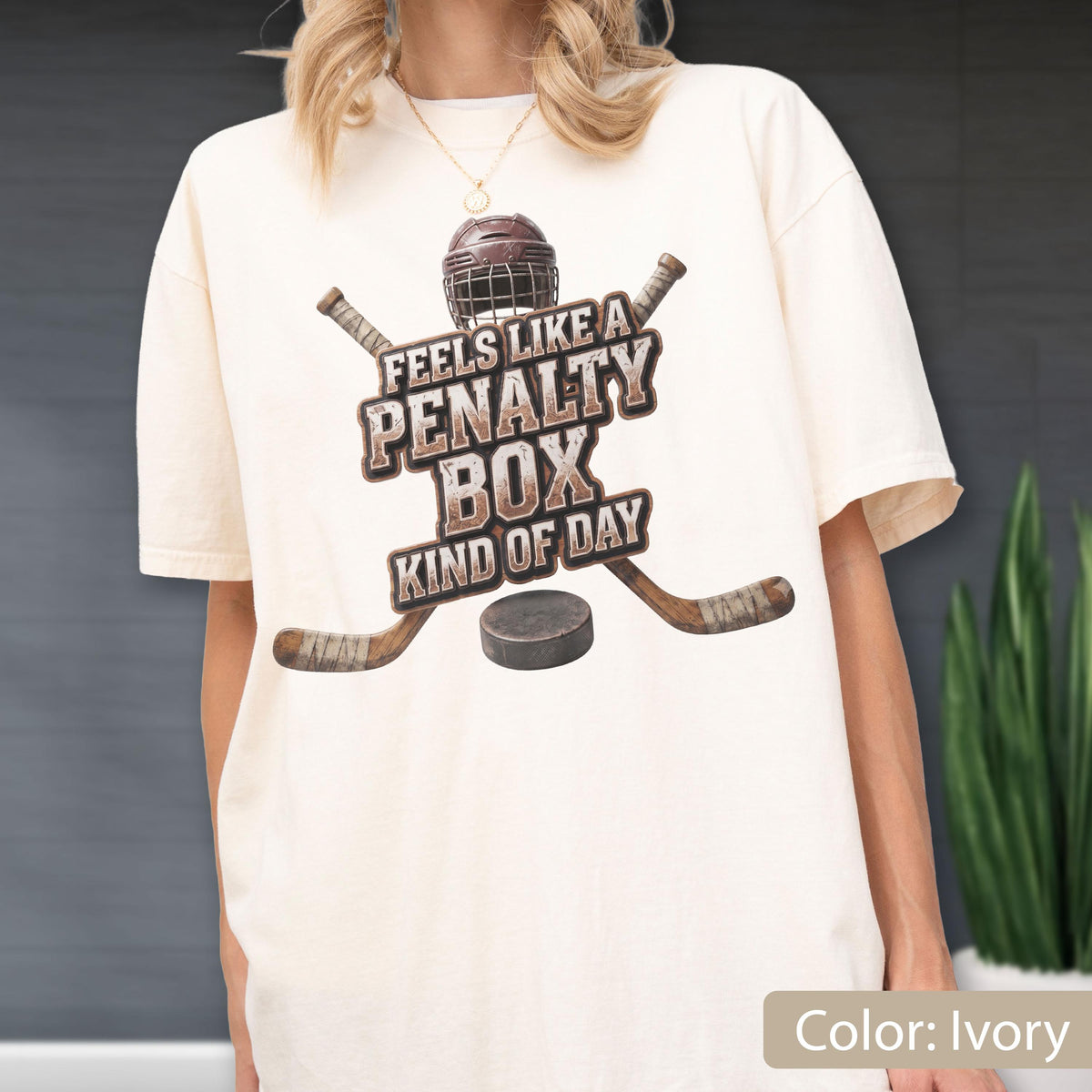 May include: An ivory short-sleeve t-shirt featuring a graphic of a brown hockey helmet, two crossed hockey sticks, and a black puck. The text reads, "FEELS LIKE A PENALTY BOX KIND OF DAY" in a distressed brown font centered on the chest area.