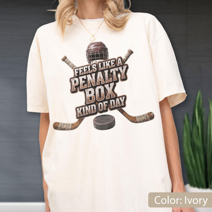 May include: An ivory short-sleeve t-shirt featuring a graphic of a brown hockey helmet, two crossed hockey sticks, and a black puck. The text reads, "FEELS LIKE A PENALTY BOX KIND OF DAY" in a distressed brown font centered on the chest area.