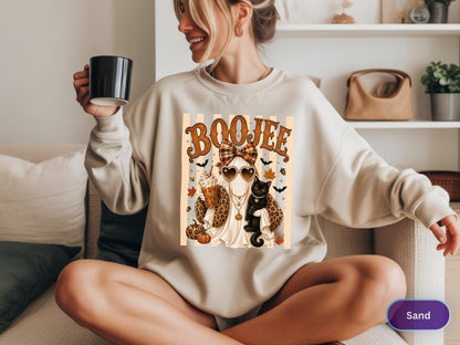 May include: A sand-colored crewneck sweatshirt featuring a graphic print of a ghost wearing a plaid bow, heart-shaped sunglasses, and a leopard print jacket. The ghost holds a coffee cup and stands next to a black cat, pumpkins, and bats.