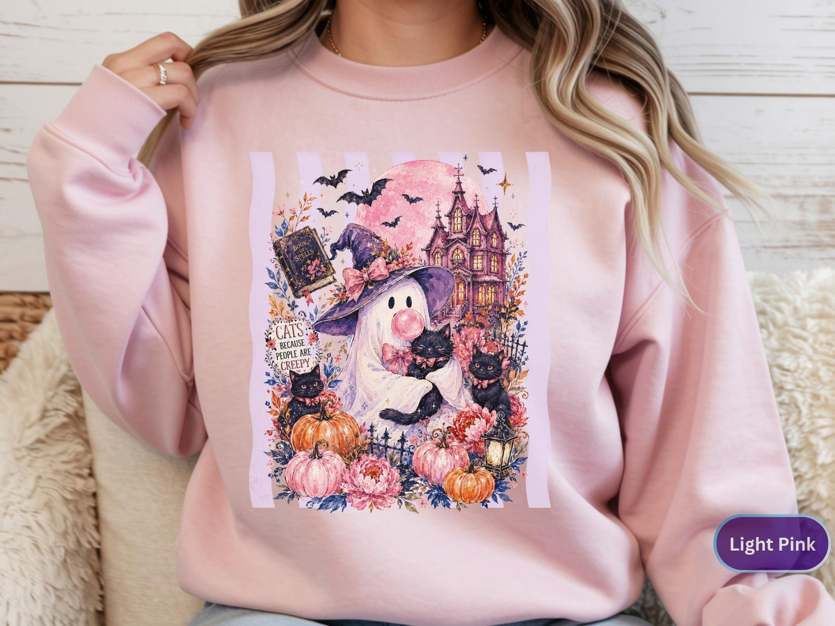 May include: A light pink crewneck sweatshirt featuring a graphic print of a ghost wearing a witch hat, holding a black cat, and blowing a bubble. The design includes pumpkins, a haunted house, bats, and the text Cats because people are creepy.