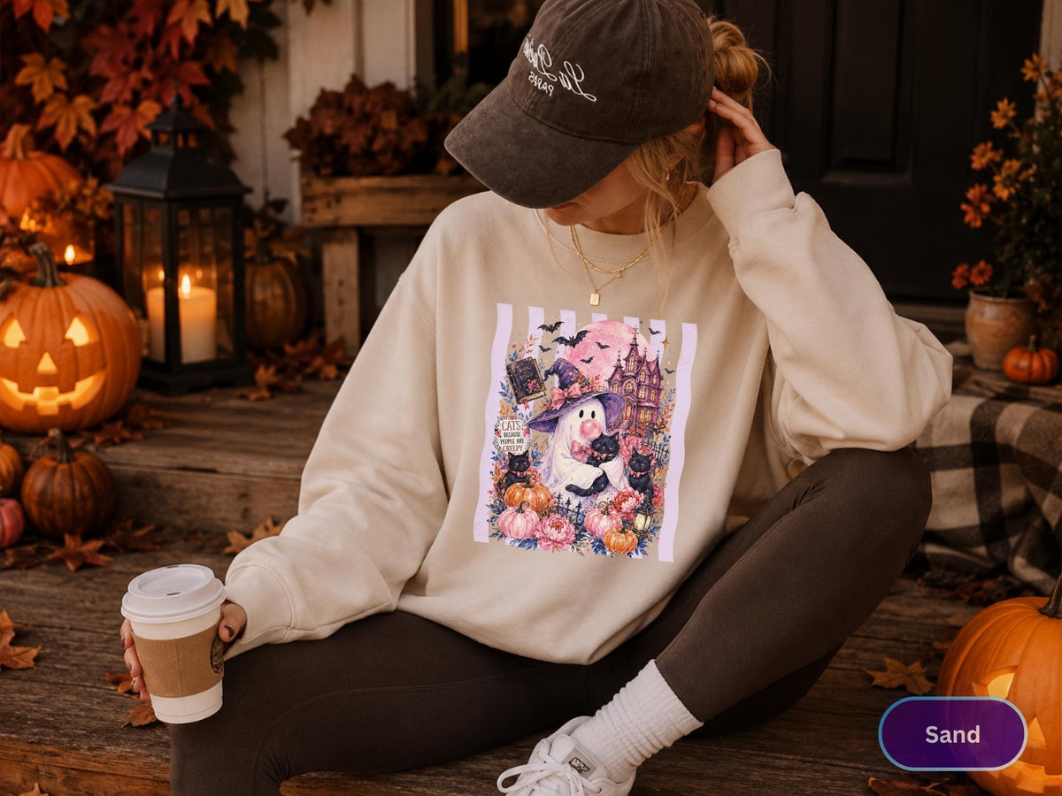 May include: A sand-colored crewneck sweatshirt featuring a graphic print of a ghost wearing a witch hat, surrounded by black cats, pumpkins, flowers, and a haunted house. The text on the graphic reads, "Eats, Shoots and Creepy". The sweatshirt is paired with dark leggings and a dark baseball cap.