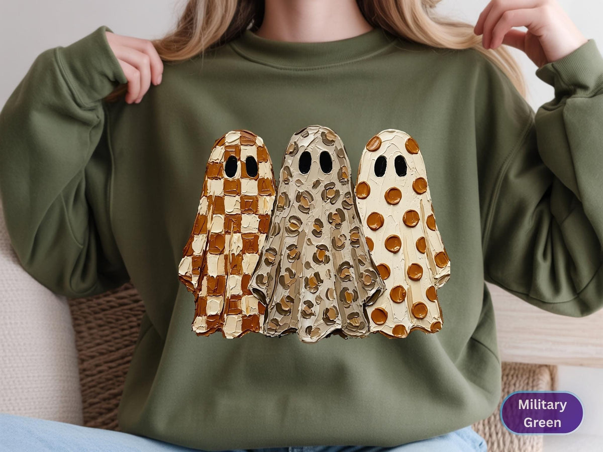 May include: A military green crewneck sweatshirt featuring a graphic of three ghosts with textured paint strokes. The left ghost has a brown and cream checkerboard pattern, the middle ghost has a leopard print, and the right ghost has brown polka dots.