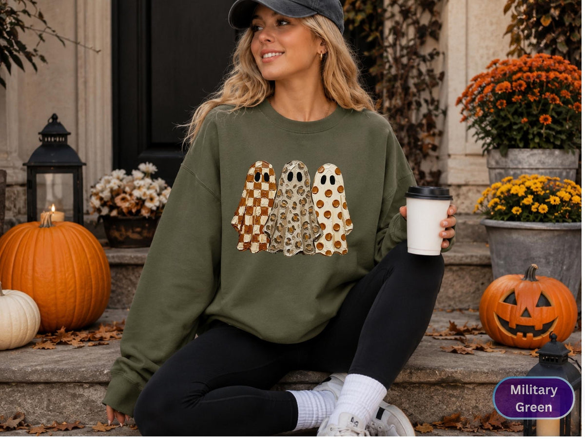 May include: A military green crewneck sweatshirt featuring a graphic print of three ghosts. The first ghost has a brown and white checkered pattern, the second has a leopard print, and the third has a brown polka dot pattern on a white background.