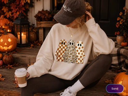 May include: A beige crewneck sweatshirt featuring a graphic print of three ghosts standing side by side. The first ghost has a brown and white checkered pattern, the second has a leopard print, and the third has a brown polka dot pattern.