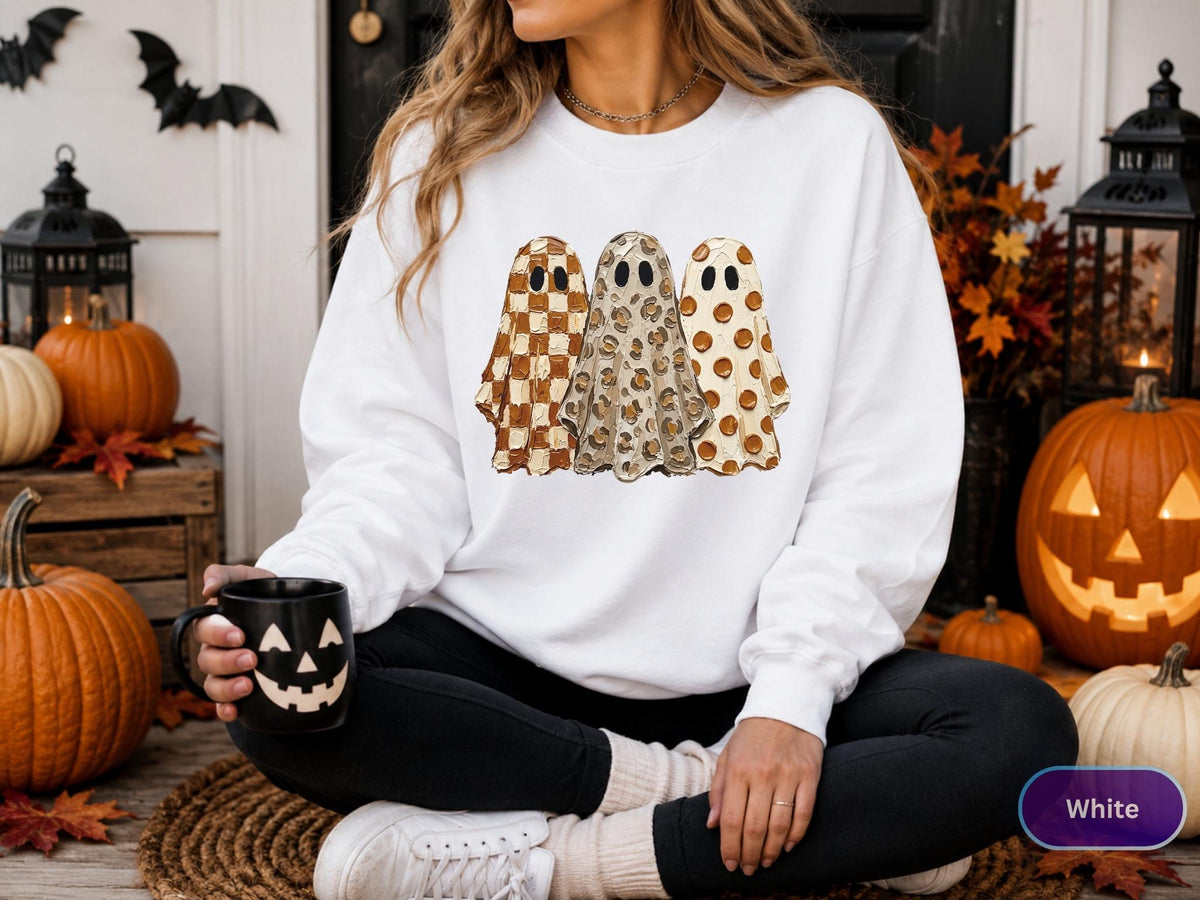 May include: A white long-sleeve sweatshirt featuring a graphic print of three ghosts standing side-by-side. The first ghost has a brown and white checkered pattern, the second has a tan leopard print, and the third has brown polka dots.