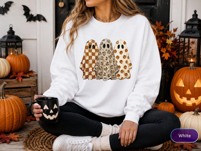 May include: A white long-sleeve sweatshirt featuring a graphic print of three ghosts standing side-by-side. The first ghost has a brown and white checkered pattern, the second has a tan leopard print, and the third has brown polka dots.