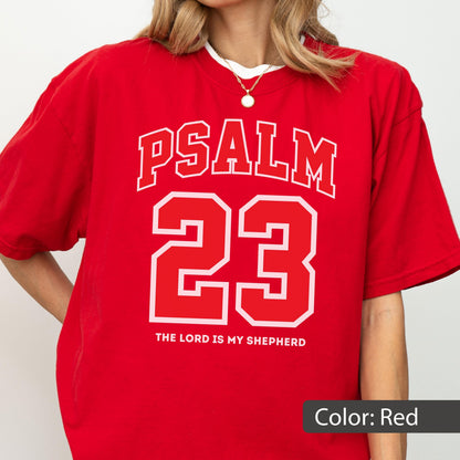 May include: A red short-sleeved t-shirt featuring white collegiate-style lettering that reads PSALM 23, with the number 23 prominently displayed below, and the text THE LORD IS MY SHEPHERD printed in a smaller white font at the bottom.