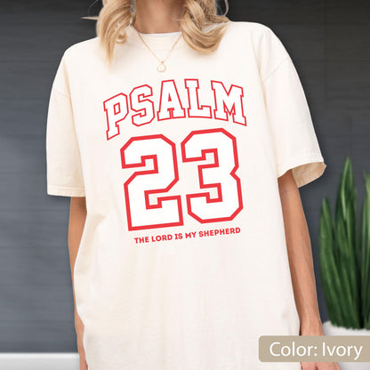May include: An ivory short-sleeved t-shirt featuring red collegiate-style lettering that reads PSALM 23 and the text THE LORD IS MY SHEPHERD below the large red number 23. The shirt has a crew neckline and a relaxed, loose-fitting silhouette.