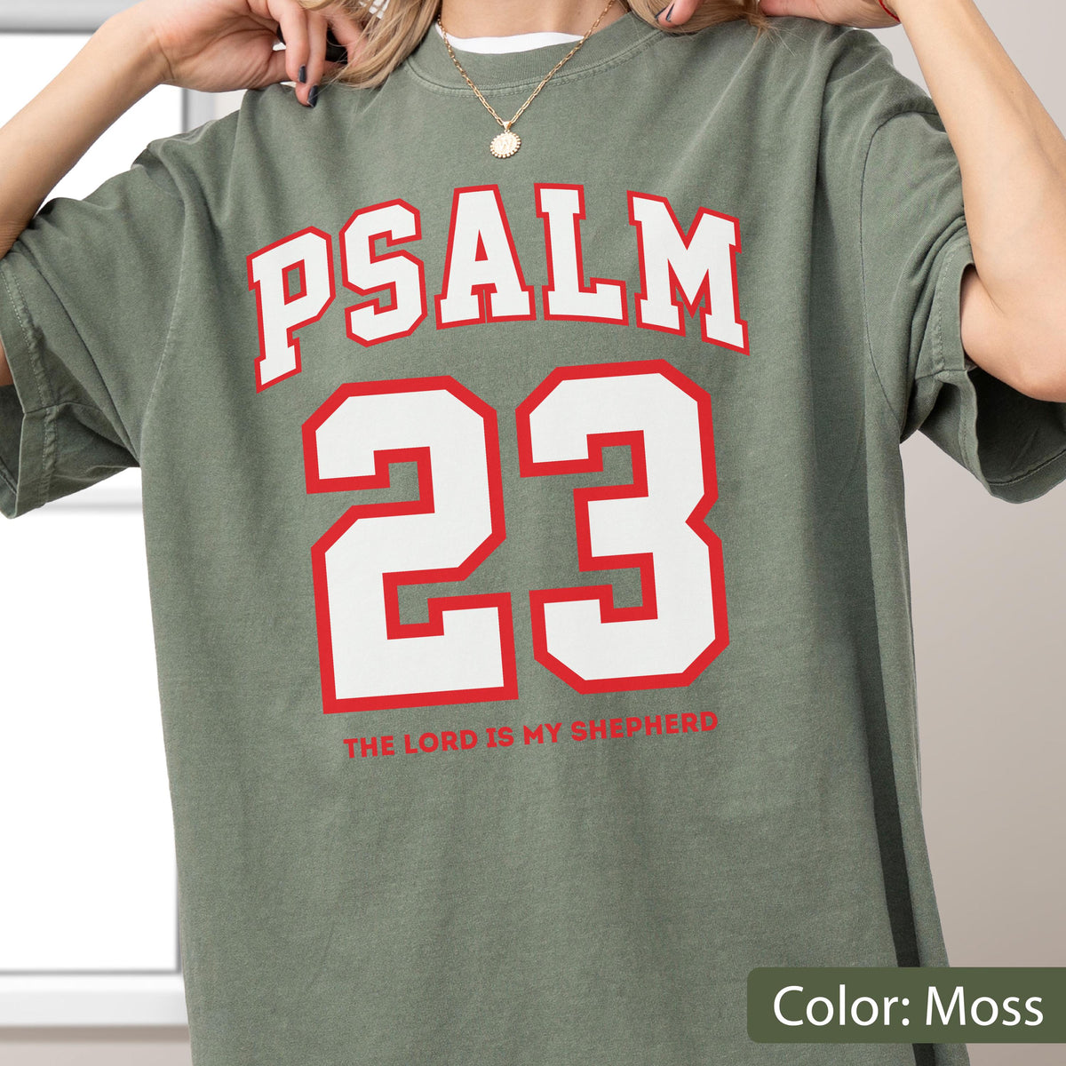 May include: A moss green short-sleeve t-shirt featuring the white text PSALM 23 in a bold arched font above a large white number 23 with a red outline. Below the number, the text THE LORD IS MY SHEPHERD is printed in a smaller red font.