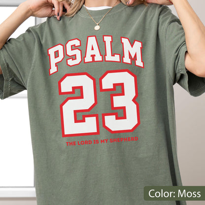 May include: A moss green short-sleeve t-shirt featuring the white text PSALM 23 in a bold arched font above a large white number 23 with a red outline. Below the number, the text THE LORD IS MY SHEPHERD is printed in a smaller red font.