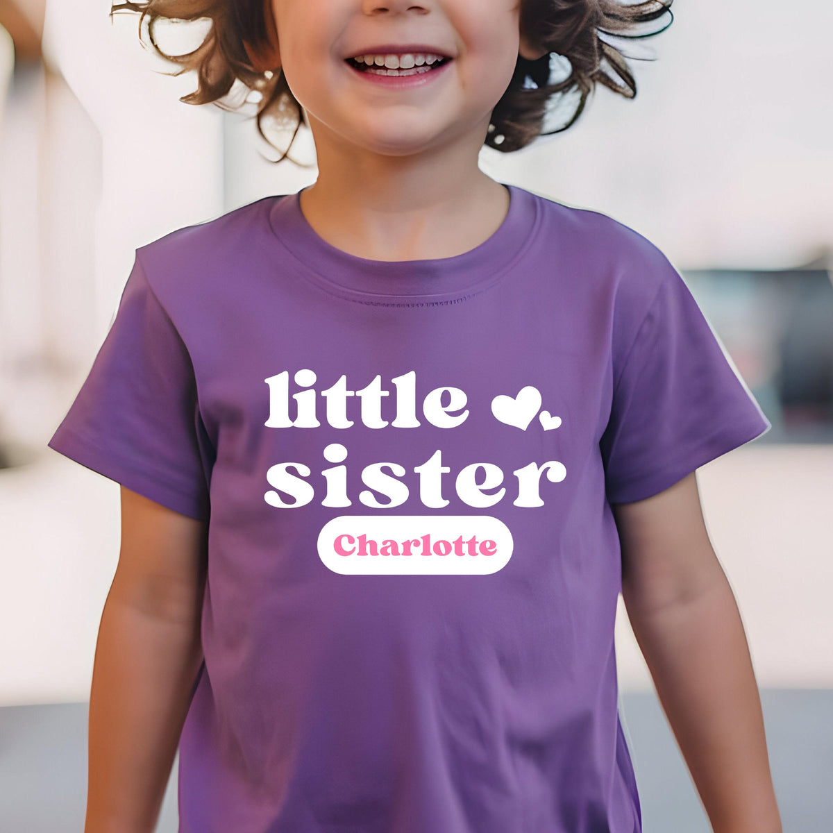 May include: A purple short-sleeved t-shirt featuring the white text "little sister" with two small white hearts. Below the text, a white horizontal oval contains the name "Charlotte" printed in a soft pink, sans-serif font.