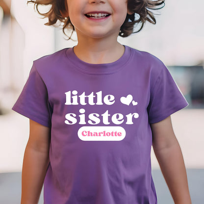 May include: A purple short-sleeved t-shirt featuring the white text "little sister" with two small white hearts. Below the text, a white horizontal oval contains the name "Charlotte" printed in a soft pink, sans-serif font.