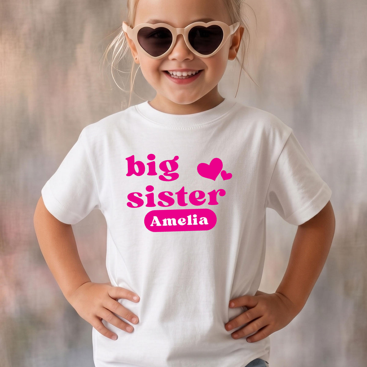 May include: A white short-sleeved t-shirt featuring the hot pink text "big sister" with two small hearts and the name "Amelia" printed inside a solid pink rounded rectangle on the chest. The shirt has a crew neckline and a standard fit.