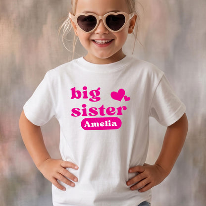 May include: A white short-sleeved t-shirt featuring the hot pink text "big sister" with two small hearts and the name "Amelia" printed inside a solid pink rounded rectangle on the chest. The shirt has a crew neckline and a standard fit.
