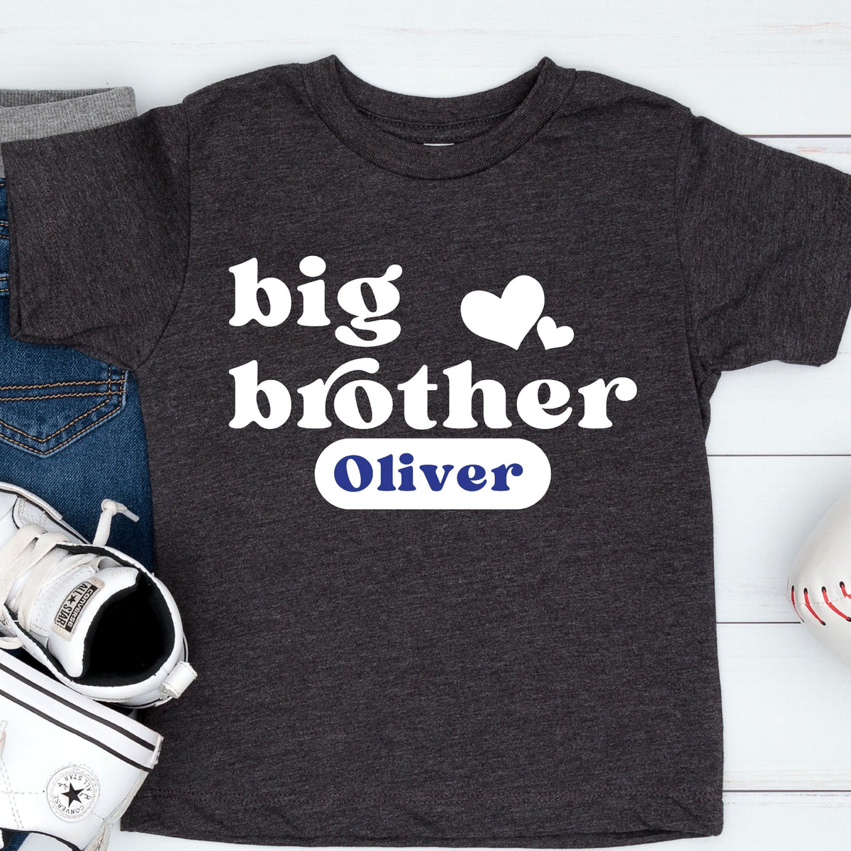 May include: A charcoal gray short-sleeve t-shirt featuring the white text "big brother" with two white heart icons above it. Below the text, a white rectangular patch displays the name "Oliver" in bold blue lettering.