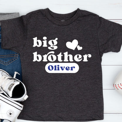 May include: A charcoal gray short-sleeve t-shirt featuring the white text "big brother" with two white heart icons above it. Below the text, a white rectangular patch displays the name "Oliver" in bold blue lettering.