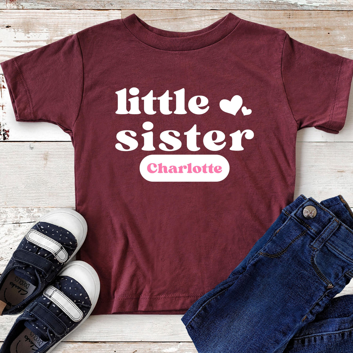 May include: A maroon short-sleeved t-shirt featuring the white text "little sister" with two small white hearts and the name "Charlotte" printed in pink inside a white rounded rectangular box, accompanied by dark blue jeans and navy sneakers.